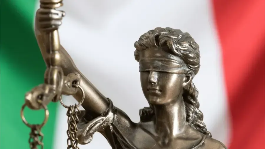 Estátua de bronze da Justiça vendada, segurando a balança, diante da bandeira italiana