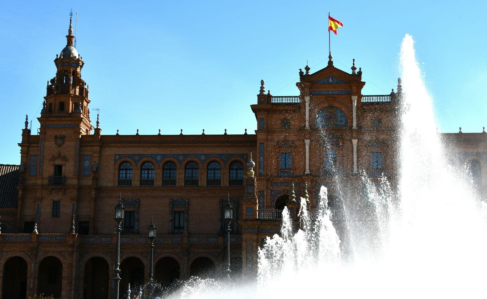 Fachada da Praça de Espanha, em Sevilha, com a bandeira espanhola no topo e o jato da fonte em primeiro plano