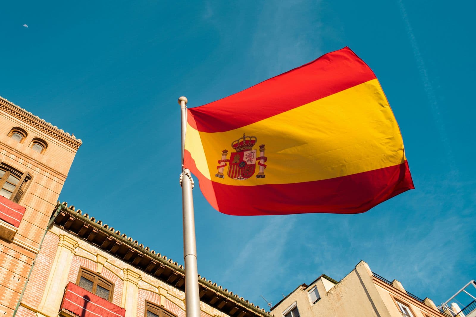 Bandeira da Espanha tremulando entre prédios antigos, vista de baixo contra o céu azul