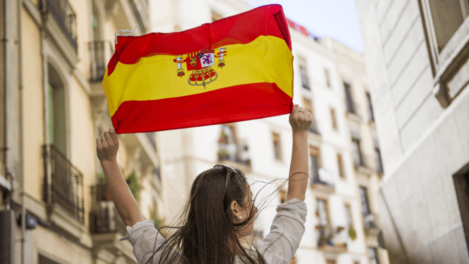 Mulher de costas erguendo uma bandeira da Espanha em uma rua estreita