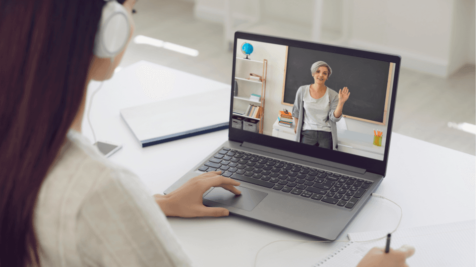 Mulher de fones assistindo a uma aula on-line pelo notebook, com a professora na tela