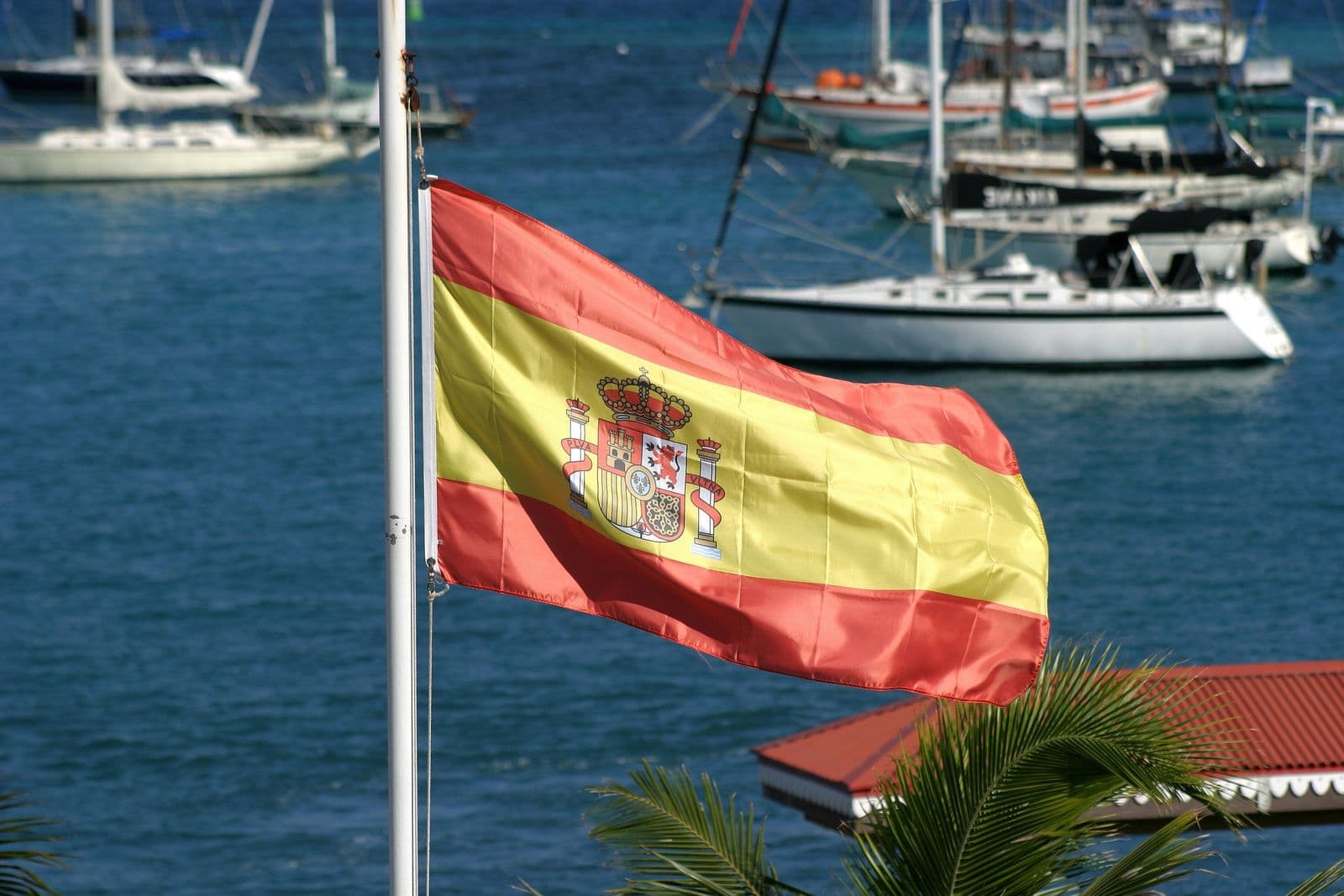 Bandeira da Espanha em um mastro à beira-mar, com veleiros ancorados ao fundo