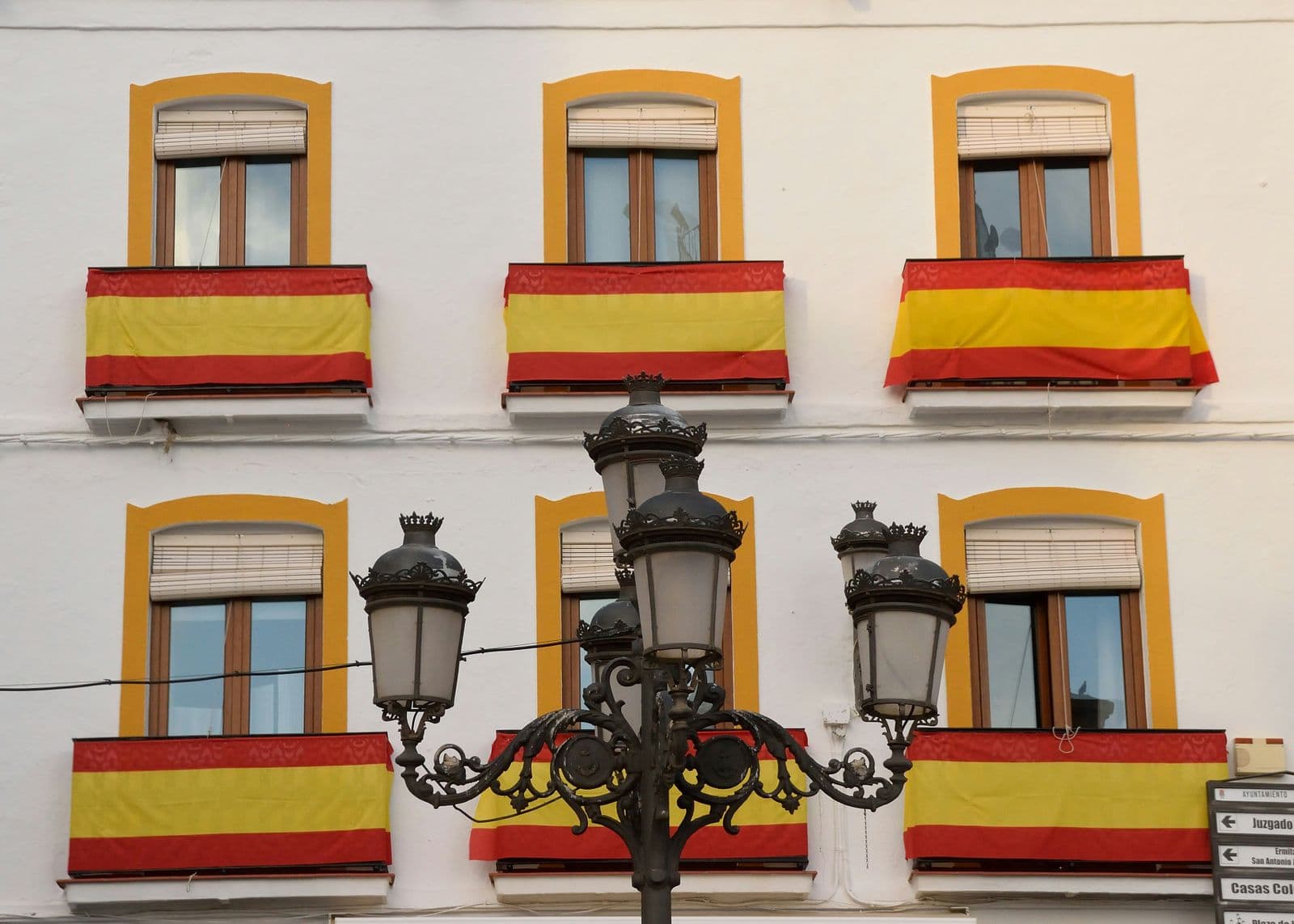 Fachada branca com bandeiras da Espanha penduradas nas sacadas e um lampião de rua em primeiro plano
