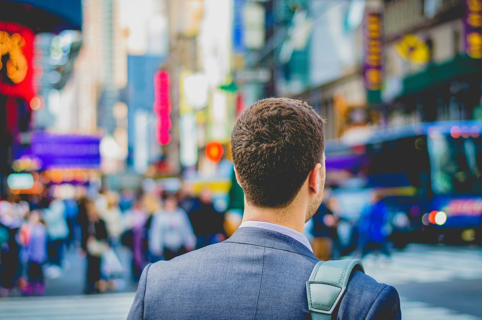 Homem de blazer e mochila, visto de costas, diante de uma avenida movimentada