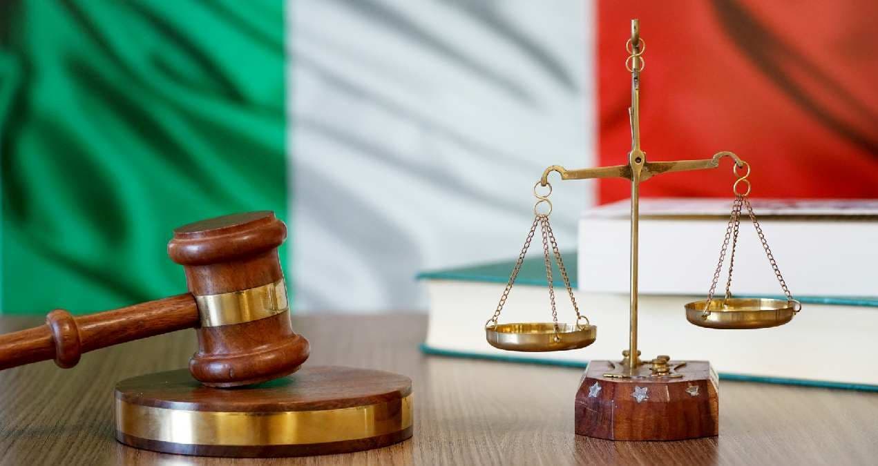 Martelo de juiz e balança dourada sobre uma mesa, diante da bandeira da Itália