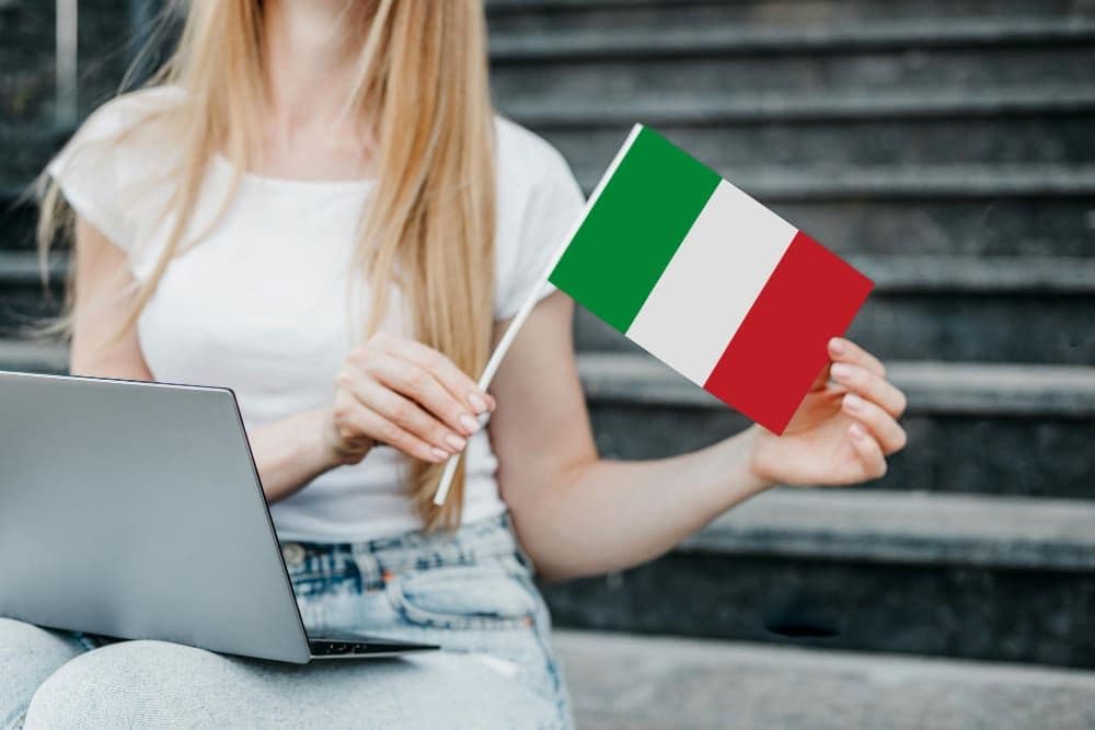 Mulher sentada em uma escada com notebook no colo, segurando uma bandeirinha da Itália
